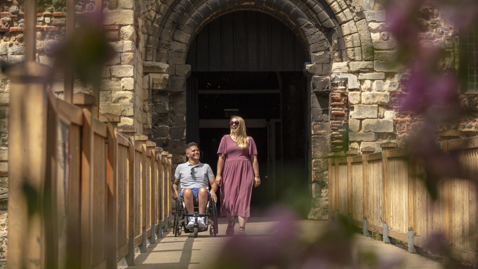 Man in wheelchair with lady, couple walking across the bridge out of Colchester Castle in Essex